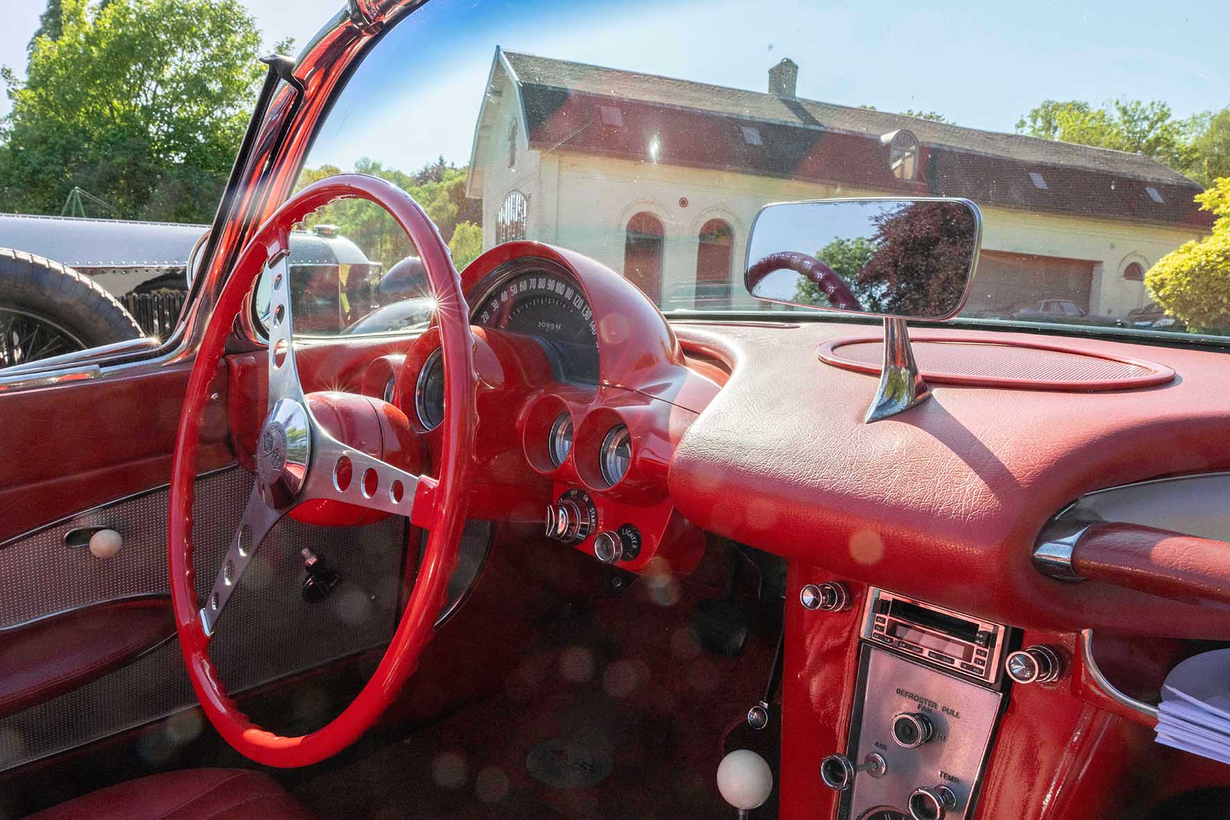 Intérieur d'une Corvette rouge décapotable des années 1960 avec volant et tableau de bord chromés, devant une maison en pierre